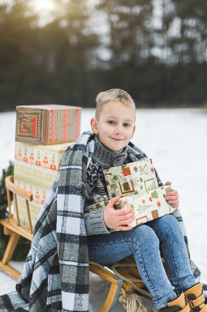 Kleiner Junge in grauem Strickpullover, bedeckt mit Plaid, sitzt auf dem Holzschlitten