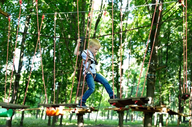 Kleiner Junge in Kletterausrüstung geht entlang einer Seilstraße in einem Abenteuerpark und hält sich an einem Seil und einem Karabiner fest.