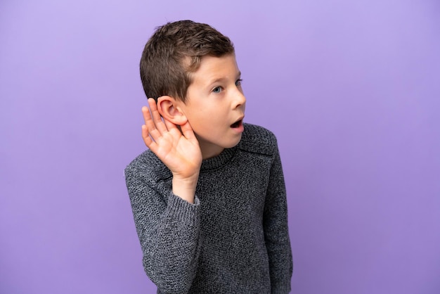 Kleiner Junge isoliert auf violettem Hintergrund, der etwas hört, indem er die Hand auf das Ohr legt