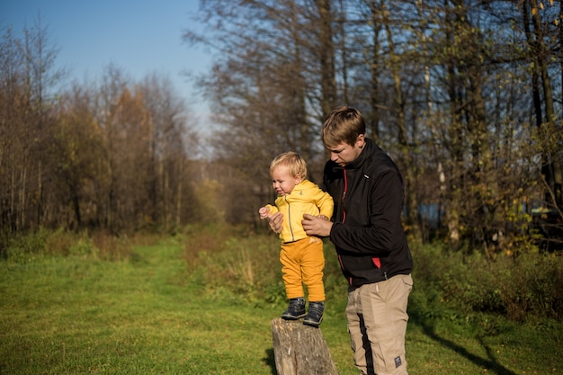 Kleiner Junge Kleinkind steht auf einem Baumstumpf, Vater unterstützt ihn vor dem Hintergrund des Waldes.