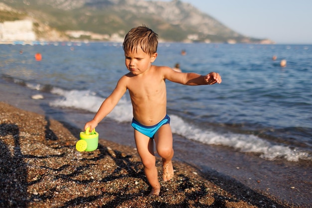 Kleiner Junge läuft mit einem Spielzeugsprinkler am sandigen Ufer entlang