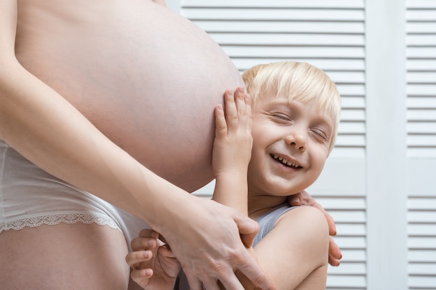 Kleiner Junge lehnte sein Ohr an den Bauch der schwangeren Mutter und lächelte. Junge schwangere Frau und ältester Sohn