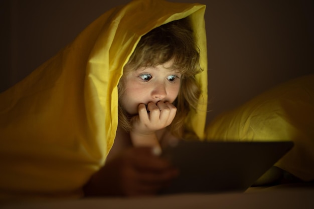 Kleiner Junge liegt auf dem Bett und surft auf einem Tablet im dunklen Raum im Internet. Kind benutzt nachts einen Tablet-PC