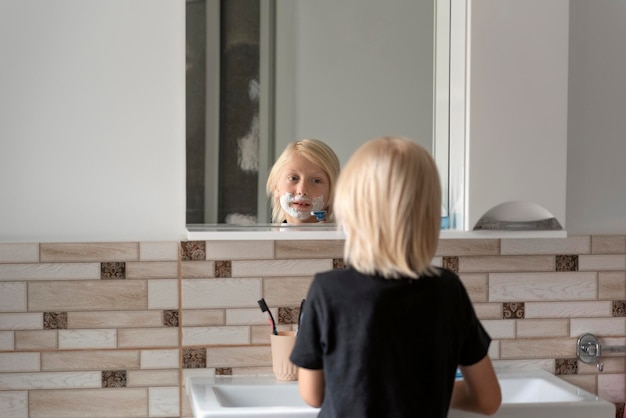 Kleiner Junge mit blonden Haaren steht vor Spiegel im Badezimmer mit Rasierschaum im Gesicht