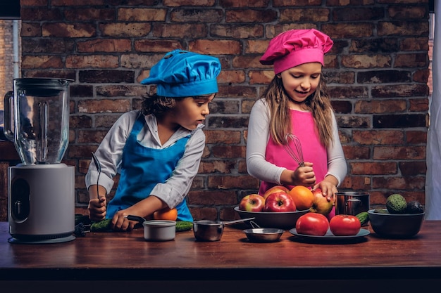 Kleiner Junge mit braunen, lockigen Haaren, gekleidet in eine blaue Kochuniform, und ein schönes Mädchen, gekleidet in eine rosafarbene Kochuniform, die zusammen in einer Küche gegen eine Ziegelwand kocht. Nettes kleines Kochpaar.