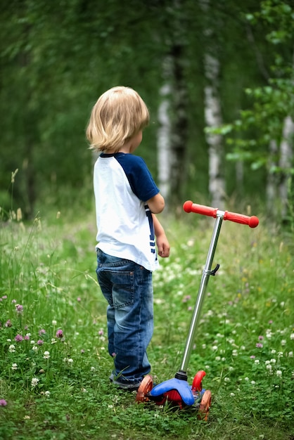 Kleiner junge mit einem roller steht auf einem grünen rasen im sommer