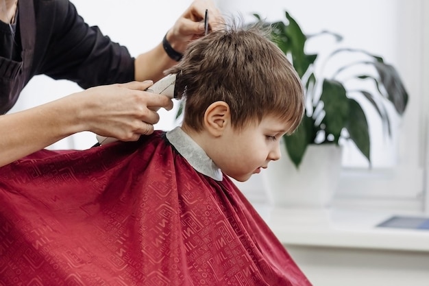 Kleiner Junge mit Friseurmaschine geschnitten Nahaufnahme von Frauenhänden, die das Haar eines Jungen im Friseursalon pflegen Beginn des Haarschnitts Porträt eines männlichen Kindes im Friseursalon, um seine Haare zu schneiden