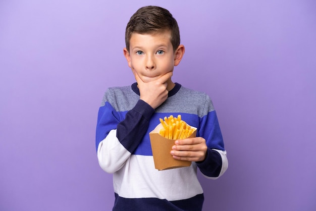 Kleiner Junge mit gebratenen Chips isoliert auf violettem Hintergrund, der Zweifel hat und nachdenkt