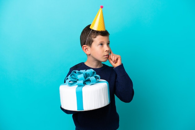 Kleiner Junge mit Geburtstagstorte isoliert auf blauem Hintergrund mit Zweifeln und Denken