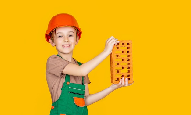 Kleiner Junge mit Helm Kinderbauhelm Schutzhelm Junge in einem Bauhelm hält einen Ziegelstein in seinen Händen auf gelbem Hintergrund Kleiner Baumeister im Helm Kind gekleidet wie ein Bauarbeiter