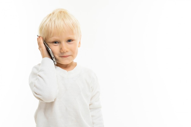 Kleiner Junge mit kurzen blonden Haaren, blauen Augen, süßem Aussehen, in weißer Jacke, hellblauer Hose, stehend und am Telefon sprechend