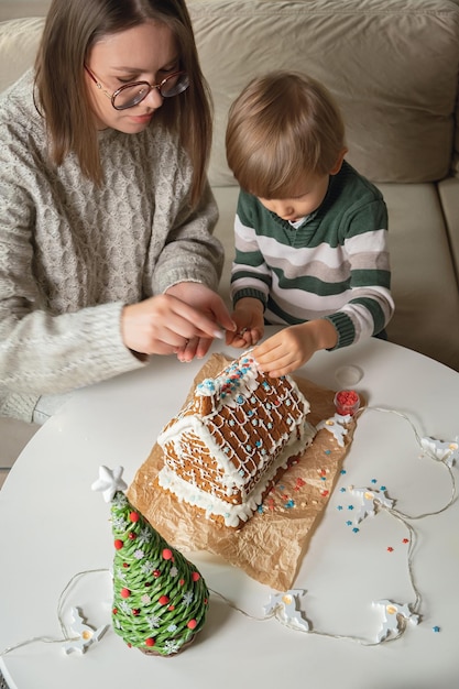 Kleiner Junge mit Mama, die zusammen Weihnachtslebkuchenhaus dekoriert, Familienaktivitäten und Traditionen an Weihnachten und Silvester