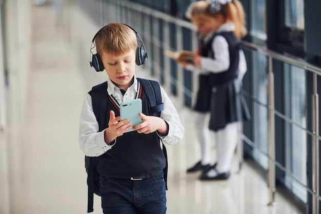 Kleiner Junge mit Telefon und Kopfhörern, die vor Schulkindern in Uniform stehen, die zusammen im Korridor stehen Konzeption der Bildung