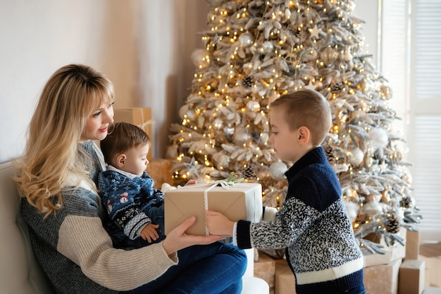 Kleiner Junge mit Weihnachtsgeschenk, der seine Mutter mit Baby in ihren Armen zu Hause grüßt. Glückliche Familie - Brüder und Mama feiern Weihnachten oder Neujahr. Frau, die auf weißer Couch nahe dem eleganten Baum sitzt.