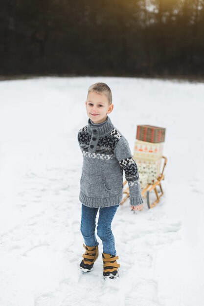 Kleiner Junge mit Weihnachtsgeschenkboxen auf Holzschlitten im Schnee