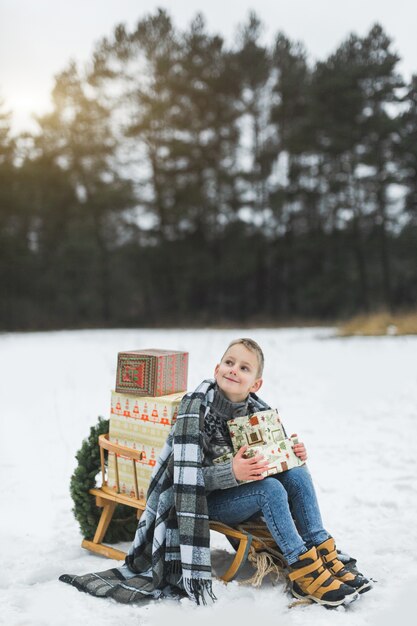 Kleiner Junge mit Weihnachtsgeschenkkästen, die auf Holzschlitten sitzen