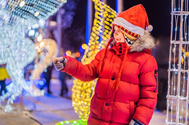 Kleiner Junge mit Wunderkerzen nahe Riesentannenbaum und Weihnachtsbeleuchtungshintergrund