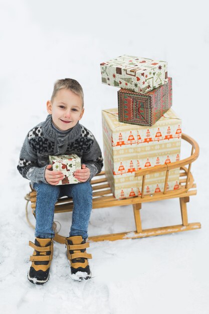 Kleiner Junge, sitzend auf dem Holzschlitten, geschmückt mit Kisten mit Weihnachtsgeschenken