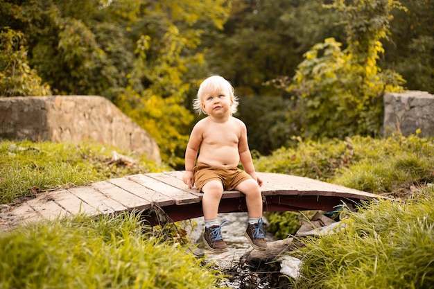 Kleiner Junge sitzt auf Brücke über dem Bach im Herbst- oder Sommerpark mit grünen und goldenen Bäumen