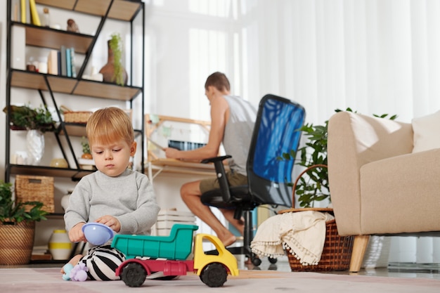 Kleiner Junge sitzt auf dem Boden und spielt mit Spielzeugauto, während sein Vater im Hintergrund am Computer arbeitet