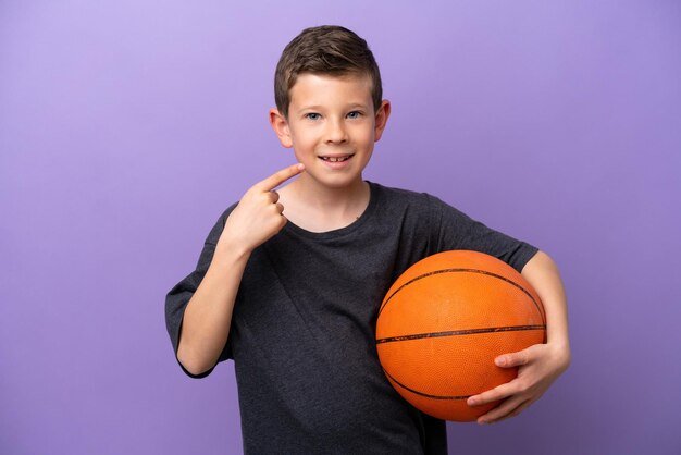 Kleiner Junge spielt Basketball isoliert auf violettem Hintergrund und gibt eine Daumen hoch Geste