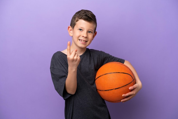 Kleiner Junge spielt Basketball isoliert auf violettem Hintergrund und macht kommende Geste