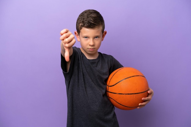 Kleiner Junge spielt Basketball isoliert auf violettem Hintergrund und zeigt Daumen nach unten mit negativem Ausdruck