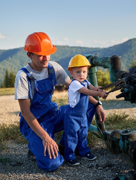 Kleiner Junge spielt mit einem Werkzeug neben einer Rohrleitung, erwachsener Mechaniker begleitet von einem Elektroingenieur im Freien, Spezialist, der Erfahrungen mit kleinen Nachkommen teilt