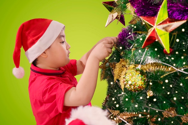Kleiner Junge trägt Weihnachtsmütze, während er zu Hause einen Weihnachtsbaum schmückt