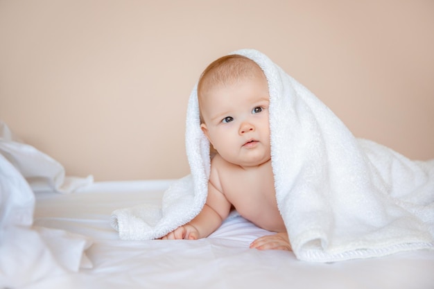 kleiner Junge zu Hause im Schlafzimmer, der nach dem Baden auf einem weißen Laken liegt, das in ein Handtuch gewickelt ist