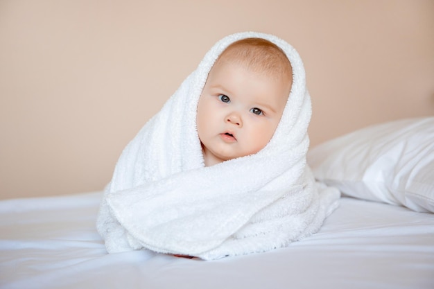 kleiner Junge zu Hause im Schlafzimmer, der nach dem Baden auf einem weißen Laken liegt, das in ein Handtuch gewickelt ist