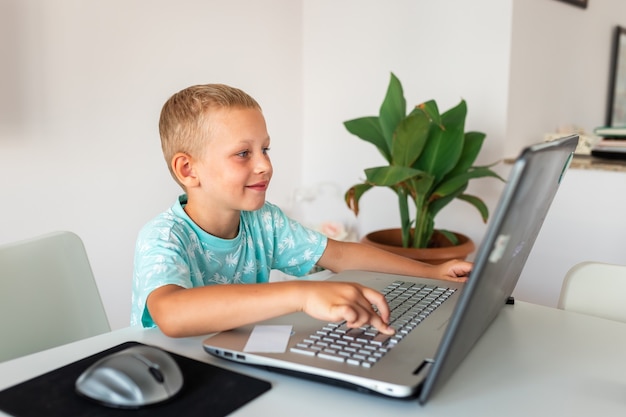 Kleiner junger Schuljunge, der zu Hause mit einem Laptop und Klassennotizen arbeitet, die in einer virtuellen Klasse studieren.