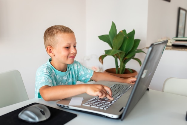 Kleiner junger Schuljunge, der zu Hause mit einem Laptop und Klassennotizen arbeitet, die in einer virtuellen Klasse studieren.