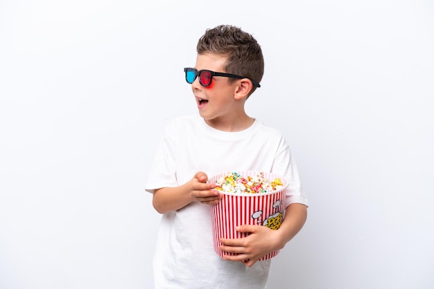 Kleiner kaukasischer Junge isoliert auf weißem Hintergrund mit 3D-Brille und hält einen großen Eimer Popcorn, während er zur Seite schaut