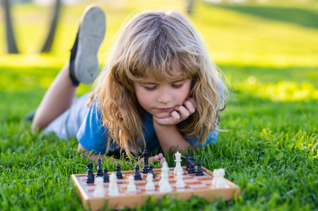 Kleiner kluger Junge, der über Schach nachdenkt Kind spielt Schach, das im Sommerpark auf Gras liegt