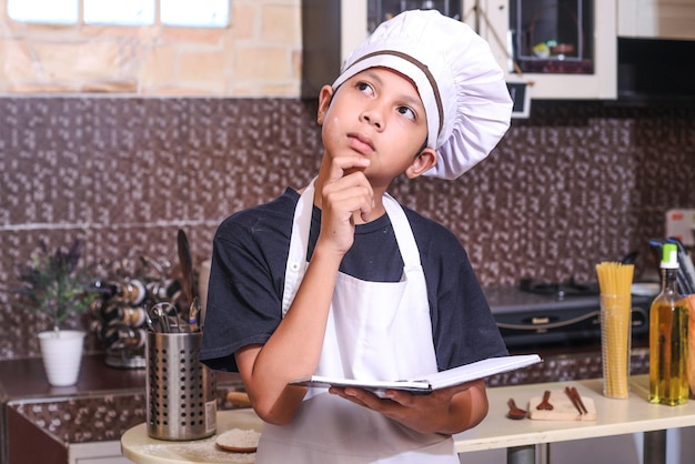 Kleiner Koch gestikuliert nachdenklich, während er zu Hause in der Küche ein Buchrezept zum Kochen in der Hand hält