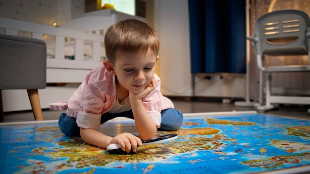 Kleiner lächelnder Junge, der im Schlafzimmer auf dem Boden liegt und durch die Lupe auf der großen Weltkarte schaut. Konzept von Reisen, Tourismus und Kindererziehung. Erkundungen und Entdeckungen für Kinder.