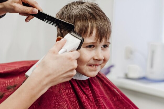 Kleiner lächelnder Junge, der mit der Friseurmaschine geschnitten wird Nahaufnahme der Frau, die die Haare des Jungen im Friseursalon pflegt Porträt des männlichen Kindes im Friseursalon, um seine Haare zu schneiden