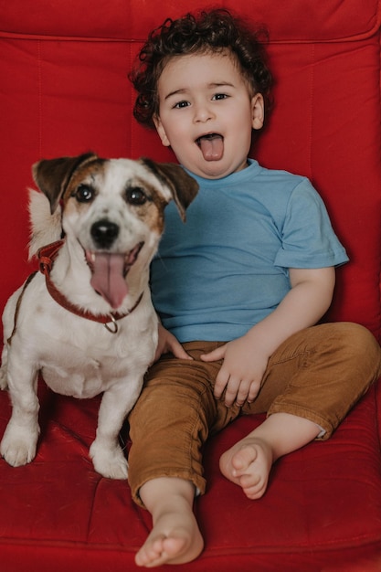 Kleiner lockiger Junge mit seinem Hund, der mit herausgestreckter Zunge auf einem roten Stuhl sitzt Lustiger Schuss von Kind und Haustier