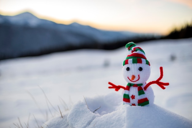 Kleiner lustiger Spielzeugbabyschneemann in gestrickter Mütze und Schal im tiefen Schnee im Freien auf unscharfem schneebedecktem Gebirgslandschaftshintergrund. Frohes neues Jahr und frohe Weihnachten Grußkartenthema.