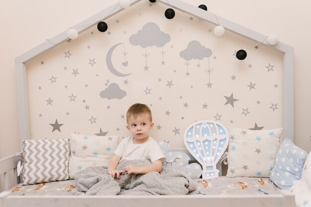 Kleiner niedlicher kleiner Junge, der im Kinderzimmer in einem hölzernen Betthaus mit Nachtlichtern in der Form eines Ballons sitzt