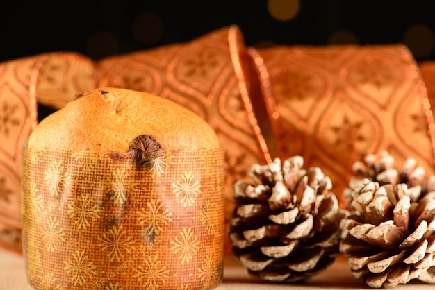 Kleiner Panettone neben Tannenzapfen und Zierband mit dunklem Hintergrund und kleinen Lichtern