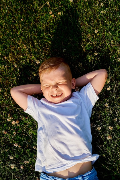 kleiner rothaariger fünfjähriger Junge liegt im Park auf dem Rücken und schielt aus der Sonnendraufsicht