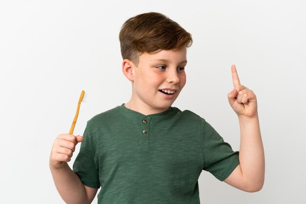 Kleiner rothaariger Junge, der eine Zahnbürste isoliert auf weißer Oberfläche hält und beabsichtigt, die Lösung zu realisieren, während er einen Finger hochhebt