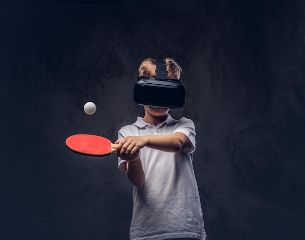 Kleiner rothaariger Junge in einem weißen T-Shirt, der mit einer Virtual-Reality-Brille Tischtennis spielt. Getrennt auf einem dunklen strukturierten Hintergrund.