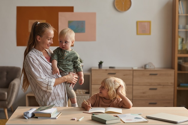 Kleiner Sohn macht Hausaufgaben am Tisch mit seiner Mutter, die ein Kind in der Hand hält und mit ihm im Zimmer spricht