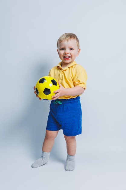 Kleiner süßer Junge, 34 Jahre alt, Fußballfan in rotem T-Shirt, der in der Hand Fußball hält, isoliert auf gelbem Hintergrund Kinder Sport Familie Freizeit Lifestyle-Konzept Werbung für den Kopierbereich