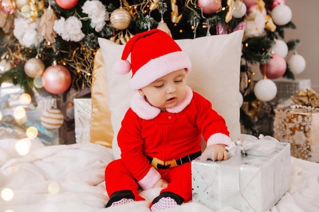 Kleiner süßer Junge im Alter von 6 Monaten, der als Weihnachtsmann verkleidet ist, sitzt in der Nähe des Weihnachtsbaums. Das Baby sitzt auf dem Boden mit Geschenken in der Nähe des Weihnachtsbaums. Weihnachten 2022