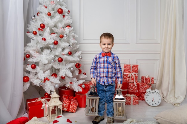 Kleiner süßer Junge, lässig gekleidet, steht im weißen Raum zwischen Weihnachtsdekorationen