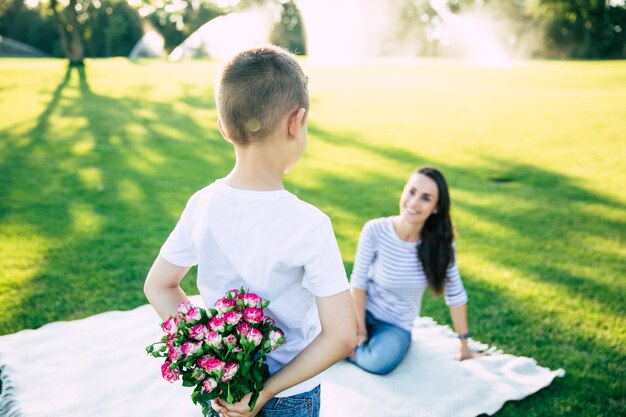 Kleiner süßer Sohn überrascht mit Blumen in den Händen für seine glückliche und schöne junge Mutter draußen im Park